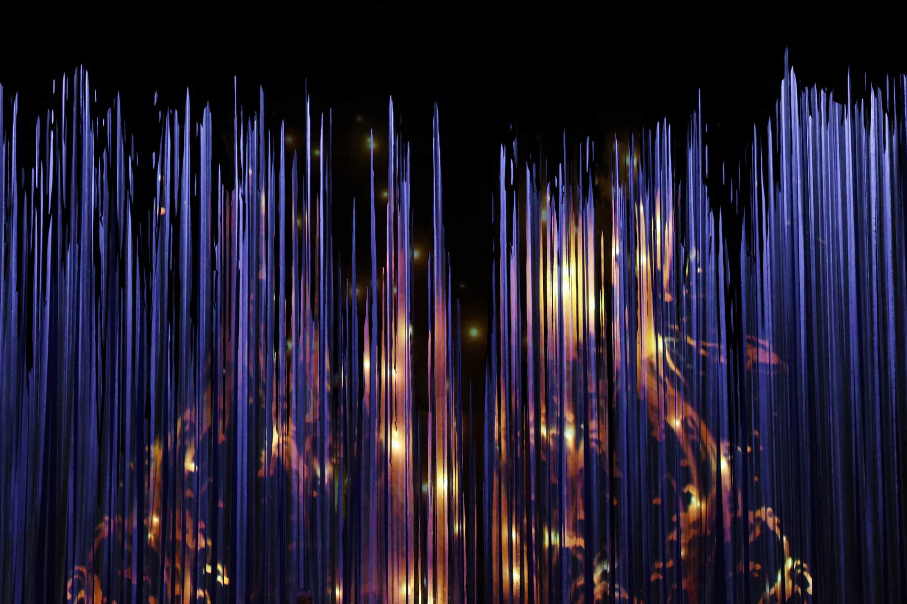 A dense hanging curtain of narrow vertical strands lit violet from above, with warm amber light burning through it from behind.