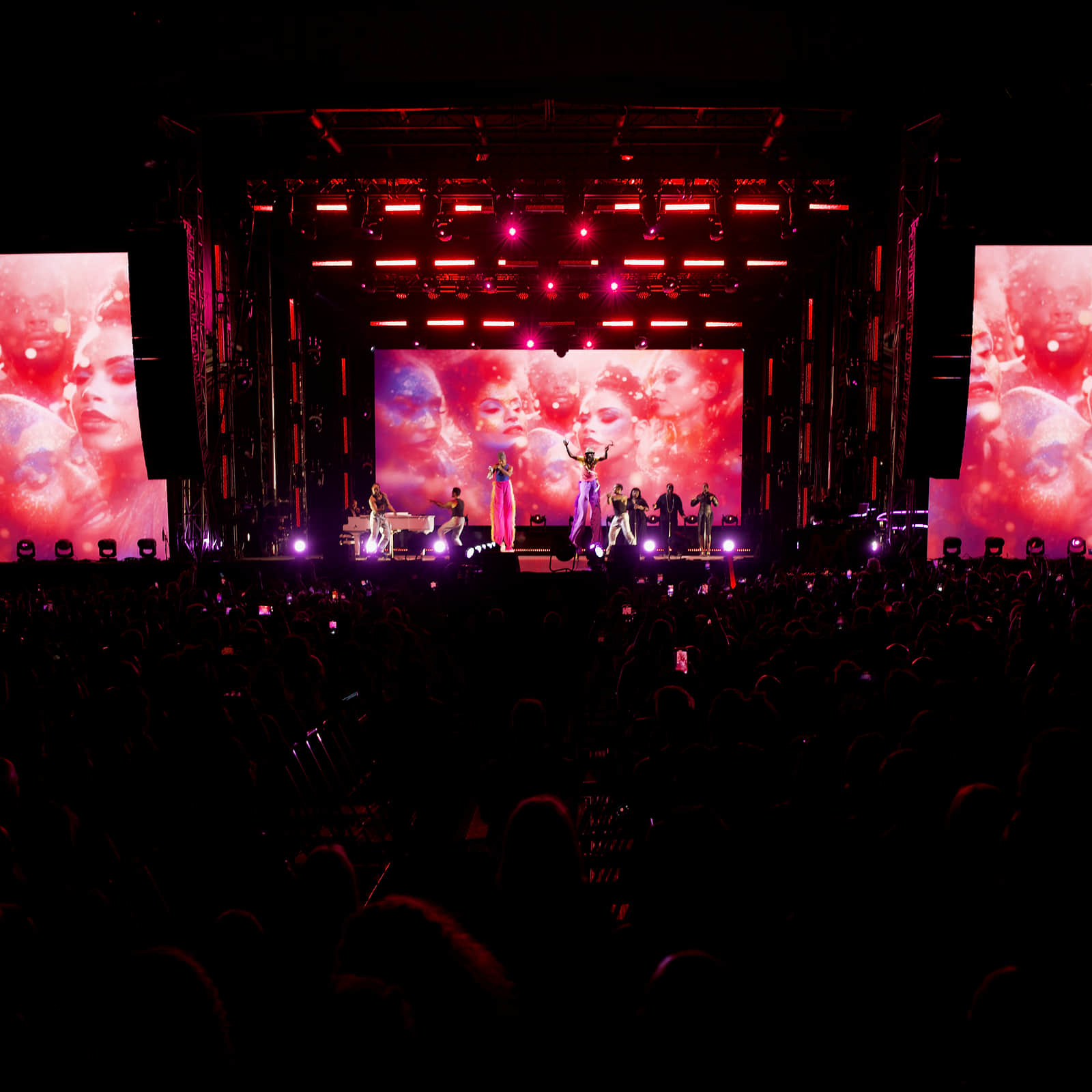 An outdoor stage seen from the crowd — a centre screen and two side screens all carrying the same pink montage of faces, performers small on the deck below, phones raised across a silhouetted audience.