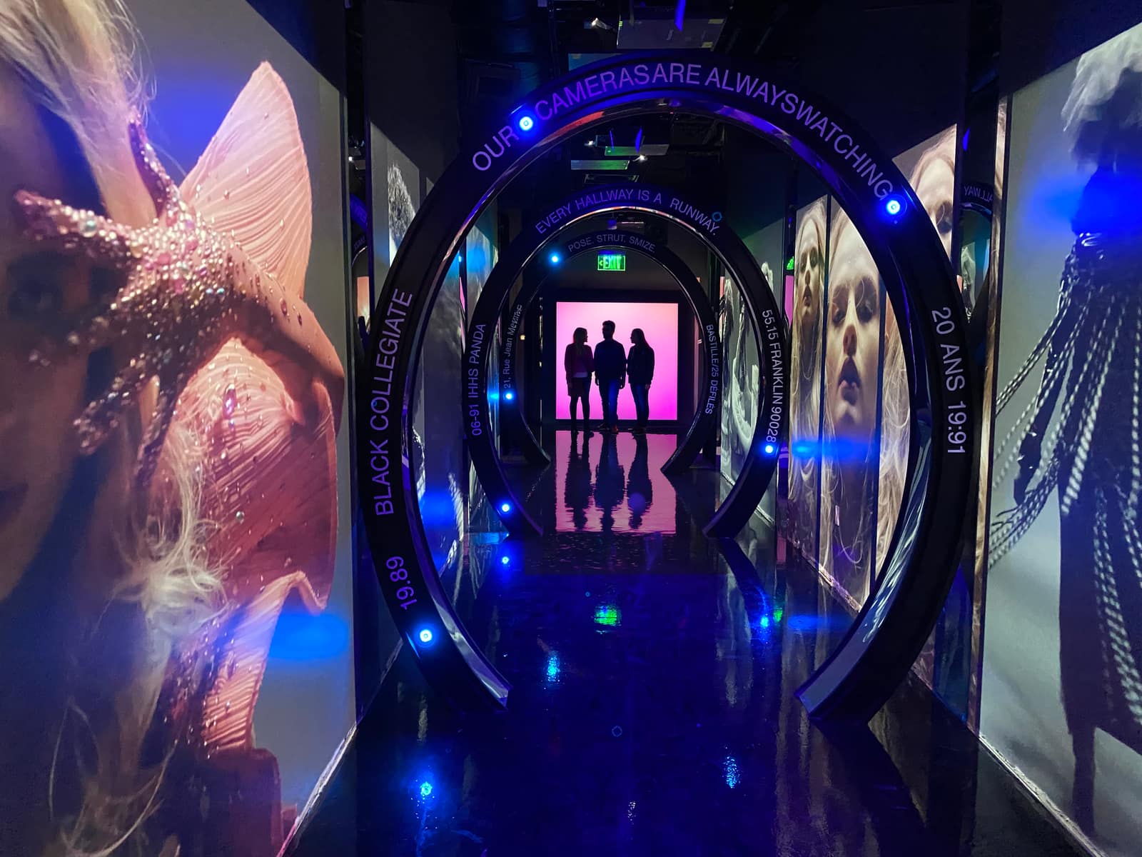 A dark corridor of illuminated elliptical arches receding to a hot-pink lightbox, three figures silhouetted at its end, backlit fashion portraits along both walls.